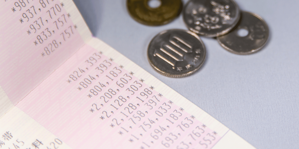 銀行の通帳が開かれ、取引金額の一覧が表示されている。通帳の隣には、穴あき5円玉や100円玉など複数の日本の硬貨が水色の背景の上に並んでいる。