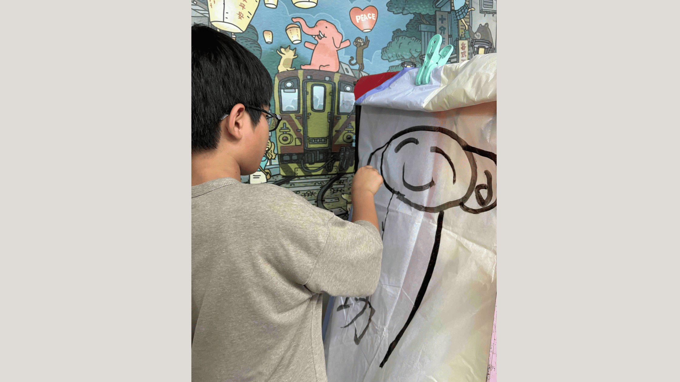 大きな白い紙に漫画風のキャラクターを描く若い人物。背景には電車や動物、ランタンなどが描かれたカラフルな壁画があり、創造的で楽しい雰囲気が漂う。